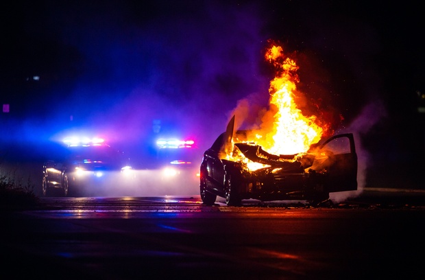 How Realistic Are Car Explosions in Movies? A totaled car sits on a road engulfed in flames. The lights of a police car are visible behind the vehicle.